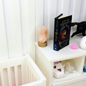 Puede incluir: Una escena de dormitorio en miniatura con una cuna blanca y una mesita de noche blanca. La mesita de noche contiene un libro titulado "Bravely" de Maggie Stiefvater, una l&aacute;mpara de sal rosa y otros objetos peque&ntilde;os. El libro tiene una cubierta azul oscuro con una mujer pelirroja.