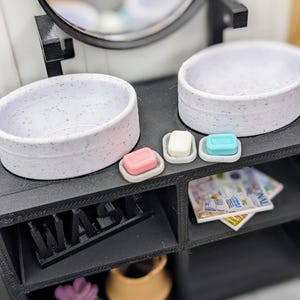 May include: Miniature bathroom set with two speckled white sinks, three soaps in pink, white, and blue, and a black vanity. The word "WASH" is displayed on the vanity. A magazine is on the shelf.