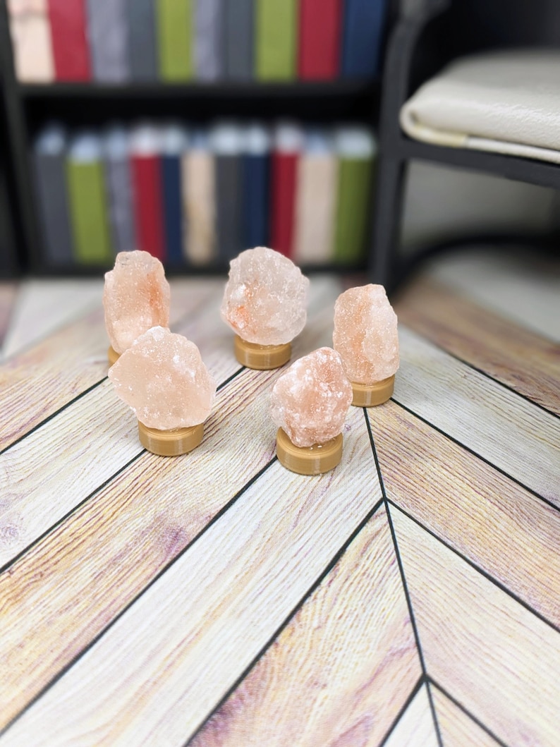 Puede incluir: Cinco l&aacute;mparas de sal rosa del Himalaya sobre peque&ntilde;as bases redondas y beige. Las l&aacute;mparas tienen una textura natural rugosa y est&aacute;n dispuestas sobre una superficie de madera clara. El fondo incluye una estanter&iacute;a con libros coloridos.