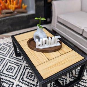 May include: Miniature coffee table with a black frame and a light wood top. A small vase with a green plant and the word "HOME" are on a brown tray. The table sits on a black and white patterned rug.