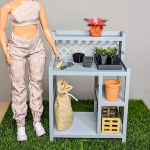 May include: A miniature gray potting bench with gardening tools, pots, and a burlap sack. The bench has a lattice back and a shelf with a small wooden crate. A doll is standing next to the bench, wearing a matching two-piece outfit.