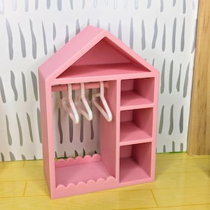 Puede incluir: Un armario rosa para casa de mu&ntilde;ecas, con forma de casa, con tejado a dos aguas. Cuenta con una barra para colgar ropa con tres perchas blancas, estantes y una base festoneada. El fondo muestra una pared con dibujos en gris claro y blanco.