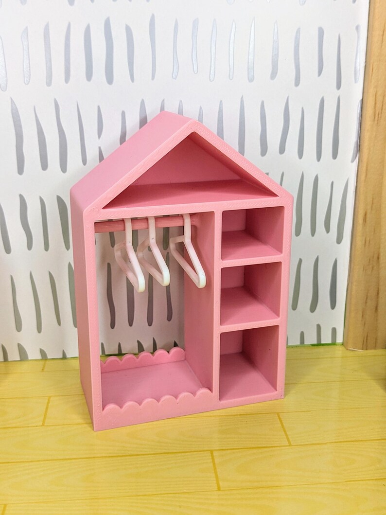 Puede incluir: Un armario rosa para casa de mu&ntilde;ecas, con forma de casa, con tejado a dos aguas. Cuenta con una barra para colgar ropa con tres perchas blancas, estantes y una base festoneada. El fondo muestra una pared con dibujos en gris claro y blanco.