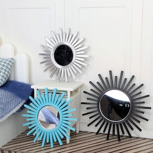 May include: Three sunburst mirrors in silver, black, and bright blue. Each mirror features a circular center glass surrounded by radiating thin, straight spokes. The silver mirror is positioned higher, while the blue and black mirrors sit lower.