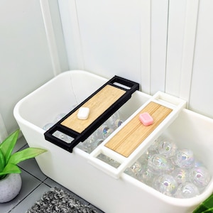 May include: A miniature white bathtub filled with iridescent bubbles. Two wooden bath trays, one black and one white, rest on the tub's edge, each holding a small bar of soap. A small potted plant and a gray and white rug are in the foreground.