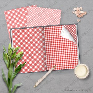 May include: A red and white gingham pattern paper, displayed open in a book with a pen and a candle on a gray surface.