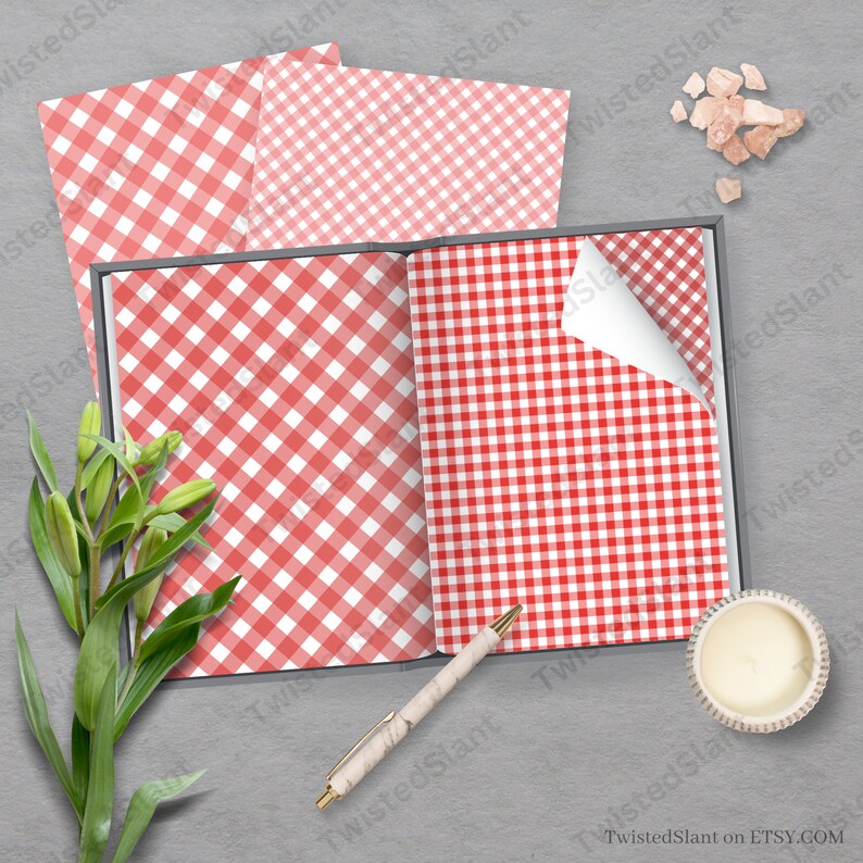 May include: A red and white gingham pattern paper, displayed open in a book with a pen and a candle on a gray surface.