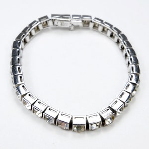 Peut inclure: Un bracelet argenté orné de pierres précieuses carrées et transparentes. Le bracelet est composé d'une série de maillons rectangulaires reliés, chacun contenant une seule pierre étincelante. Le fermoir est de conception simple et sûre. Le bijou est posé sur fond blanc.