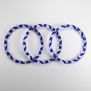 May include: Three transparent glass rings with a blue and white striped pattern, intertwined. The rings have a twisted, rope-like design. The background is plain white.