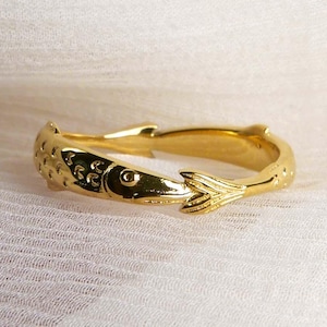 Könnte beinhalten: Ein goldfarbener Ring mit Fischdesign. Der Ring zeigt einen detaillierten Fisch mit Schuppen, einem Auge und einem Schwanz. Der Ring ist vor einem weichen, weißen Hintergrund platziert.