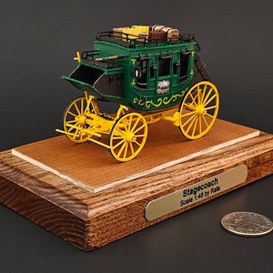 May include: A green and yellow stagecoach model on a wooden base. The model is 1:48 scale and is made by Rada. The base has a brass plaque that reads "Stagecoach Scale 1:48 by Rada".