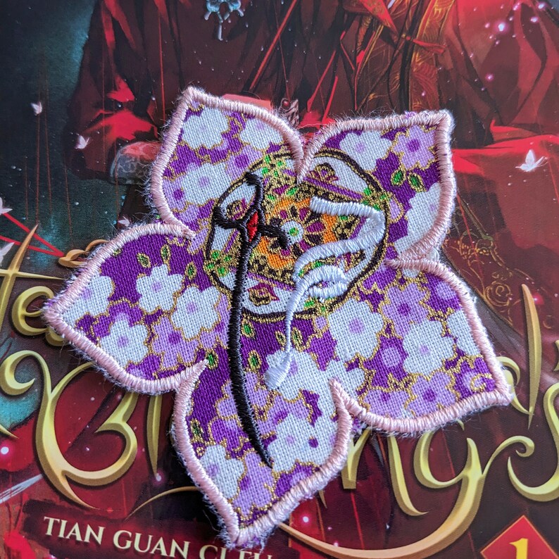 May include: Embroidered floral patch in shades of purple, white, and gold. The patch features a stylized design with a central emblem and a delicate pink border. The patch is placed on a book cover with gold lettering.