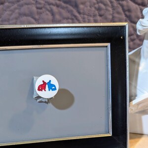 May include: A white round button with a red and blue silhouette of two rabbits facing each other. The button is attached to a black picture frame.