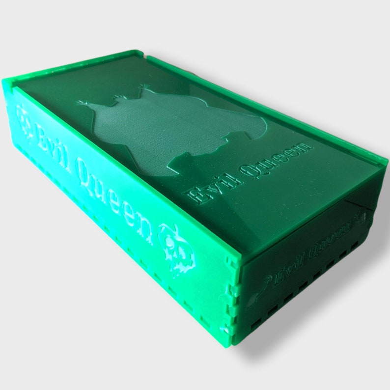 Pu&ograve; includere: Una scatola di plastica verde con le parole "Evil Queen" e "Pixel Queen" stampate su di essa. La scatola ha un design in rilievo di una silhouette di una regina.