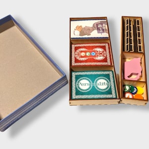 Puede incluir: Un organizador de juegos de madera con múltiples compartimentos. El organizador contiene cartas de juego con ilustraciones y texto, piezas de juego y una pieza de juego rosa. El organizador está junto a una caja con borde azul.