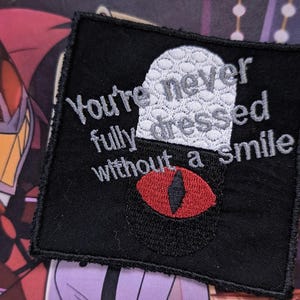May include: Black embroidered patch with white text that reads "You're never fully dressed without a smile" and a red eye with a black pupil.