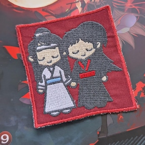 May include: Embroidered patch featuring two cartoon figures, one in white and one in black, standing side-by-side on a red background.