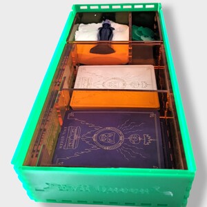Pu&ograve; includere: Un organizzatore in acrilico verde con divisori trasparenti per riporre pezzi di gioco e carte. L'organizzatore ha la scritta "Evil Queen" stampata sul davanti.