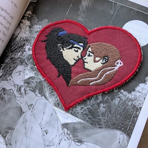 May include: Embroidered patch in the shape of a red heart with two figures inside. The figures are a man and a woman with brown hair, facing each other. The man is on the left and has blue eyes. The woman is on the right and has brown eyes. The figures are embroidered in brown thread. The heart is embroidered in red thread.
