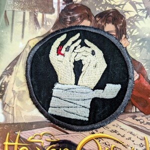 May include: Black and white embroidered patch with two hands clasped together, one hand has a red ring on the finger. The hands are wrapped in white bandages. The patch is circular and has a black border.