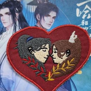 May include: A red heart-shaped embroidered patch with two anime-style characters, one with dark hair and the other with brown hair, looking at each other. The characters are surrounded by red and gold leaves.