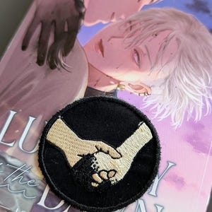 May include: Black embroidered patch with two hands clasped together. The patch is round and has a black border.