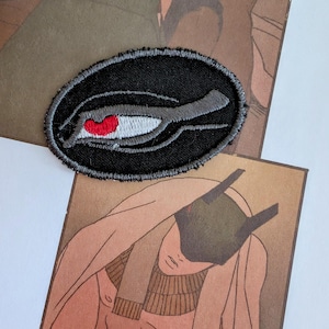 May include: Black embroidered patch with a stylized eye design. The eye is outlined in gray and has a red iris.