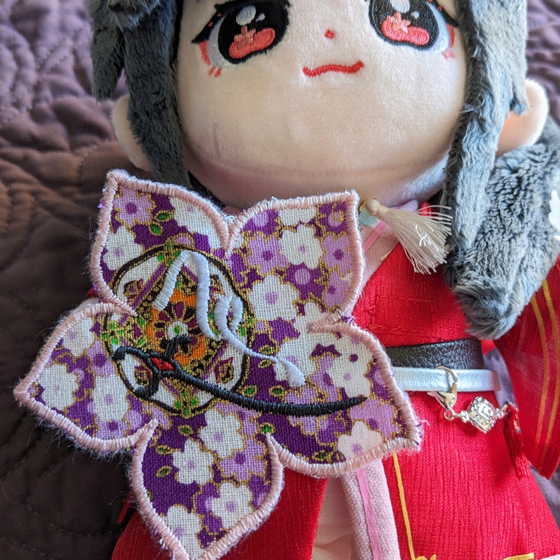 May include: A plush doll with a flower-shaped patch. The patch features a purple floral pattern with gold accents and white embroidery. The doll is dressed in a red outfit with a decorative belt and a gray fur-trimmed hat.
