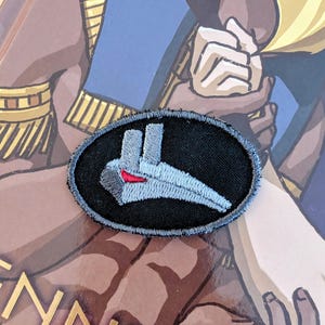 May include: A black and gray embroidered patch with a gray spaceship design. The spaceship has a red stripe on the front.
