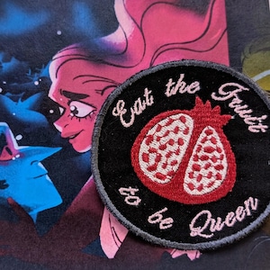 May include: A black and gray circular embroidered patch with pink stitching. The patch features a red pomegranate with white seeds and the text "Eat the Fruit to be Queen".