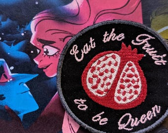 May include: A black and gray circular embroidered patch with pink stitching. The patch features a red pomegranate with white seeds and the text "Eat the Fruit to be Queen".
