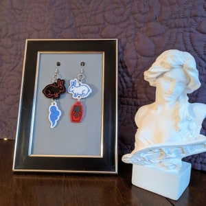 May include: A pair of dangle earrings featuring a black rabbit and a white rabbit, each with a matching charm. The charms are a blue swirl and a red bottle. The earrings are displayed in a black framed picture.