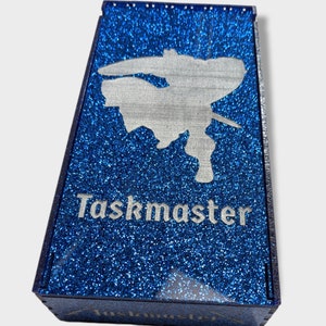 May include: A blue glitter-covered box with the word "Taskmaster" in silver lettering. A silver silhouette of a person is above the text. The box is rectangular and has a lid.