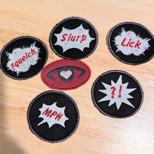 May include: Six embroidered patches with various designs and text. The patches are black with white and red embroidery. The designs include a heart with an eye, a speech bubble with the word "slurp", a speech bubble with the word "lick", a speech bubble with the word "squelch", a speech bubble with the word "mph", and a speech bubble with the text "?!".