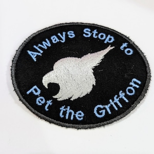 May include: A black and white embroidered patch with a white griffin silhouette and blue text that reads "Always Stop to Pet the Griffin".