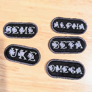 May include: Five black and white oval patches with white lettering and purple floral designs. The patches read "Alpha", "Beta", "Omega", "Uke", and "Seme".