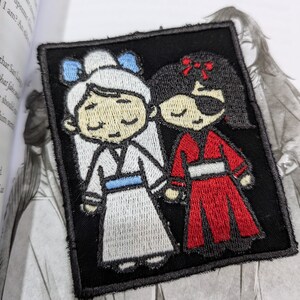 May include: Embroidered patch featuring two cartoon figures, one in a white dress with blue accents and the other in a red dress with a black eye patch. The patch is set against a black background.