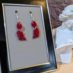 May include: A pair of red dangle earrings featuring a crab design. The earrings are hanging from silver hooks.