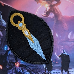 May include: A black embroidered patch with a gold and blue dagger design. The dagger has a circular handle with a blue gem in the center.