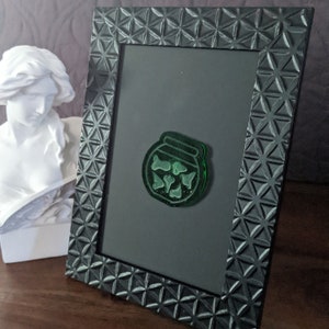 May include: A green glass jar with a lid, filled with small green bones, is displayed in a black picture frame with a geometric pattern.
