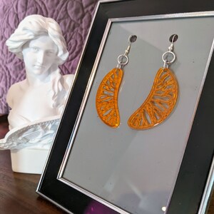 May include: A pair of orange, citrus-slice-shaped earrings with silver-colored hooks. The earrings are displayed in a black-framed picture. A white bust of a woman is in the background.