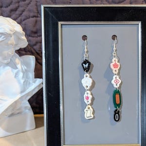 May include: A pair of dangle earrings with charms. The left earring has a black butterfly, a white flower, a pink flower, and a white scroll. The right earring has a pink maple leaf, a white eye, a green rectangle, and a black zero.
