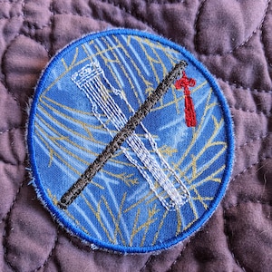 May include: A round, embroidered patch featuring a traditional Chinese instrument, the guqin, in white thread against a blue background with gold accents. A black diagonal line and a red tassel add detail. The patch has a blue border.