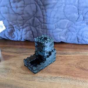 May include: A black, 3D-printed dice tower, resembling a castle tower with crenellations. The tower has a rectangular base and a taller, square upper section. The surface has a brick-like texture. The item is on a wooden surface.