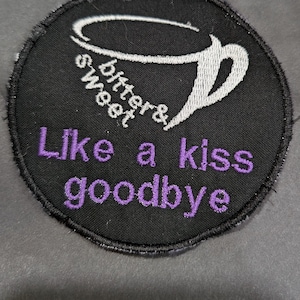 May include: A black round patch with white embroidery of a coffee cup and the text "bitters & sweet" around the cup. The text "Like a kiss goodbye" is embroidered in purple below the cup.