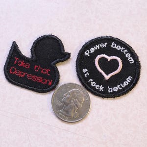 May include: Two black embroidered iron-on patches. One patch is shaped like a duck and says "Take that Depression". The other patch is round and says "Rawr bottom of rock bottom" with a pink heart in the center.