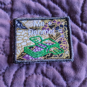 May include: Embroidered patch with the text "Me & Dammel" in white script. The patch features a green and purple design of a box with a stylized animal. The patch has a gold, purple, and black patterned background and a gray border.