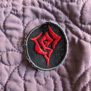 May include: A black and red embroidered patch with a stylized symbol. The symbol is a red, stylized letter "D" with a smaller red circle inside the letter.