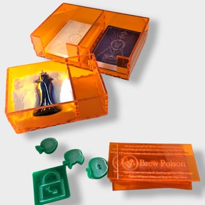 Pu&ograve; includere: Scatole di immagazzinaggio in plastica arancione con scomparti per pezzi di gioco e carte. Le scatole contengono una statuetta in miniatura, una carta con il testo "Evil Queen" e una carta con il testo "Brew Poison".