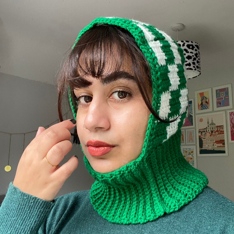 Pode incluir: Um chap&eacute;u de croch&ecirc; verde e branco com capuz. O chap&eacute;u &eacute; feito com um padr&atilde;o xadrez e tem um cachecol de pesco&ccedil;o grosso e quente.